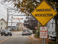 Sign reading Dangerous Intersection as cars navigate intersection in background.